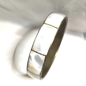 Könnte beinhalten: Ein silberfarbenes Armreif-Armband mit einem rechteckigen, segmentierten Design. Die Segmente bestehen aus schillerndem weißem Material, möglicherweise Perlmutt, und sind durch dünne, dunkle Linien getrennt. Das Armband hat eine glatte, polierte Oberfläche.