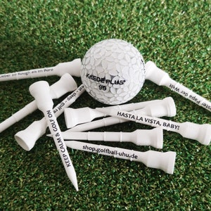 Könnte beinhalten: Ein weißer Golfball mit einem weißen und grauen Muster. Der Ball ist von acht weißen Golftees mit verschiedenen Sprüchen umgeben, darunter "Keep Calm & Golf On", "Hasta La Vista, Baby!" und "Lieber im Rough als im Büro".