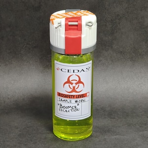 Puede incluir: Un vial cilíndrico transparente lleno de un líquido amarillo neón brillante. La tapa blanca cuenta con un mecanismo de bloqueo rojo y un pasador de metal. Una etiqueta blanca muestra el logotipo de CEDA, un símbolo de riesgo biológico y el texto: BIOSAFETY LEVEL, SAMPLE #0841, BOOMER EXCRETION.