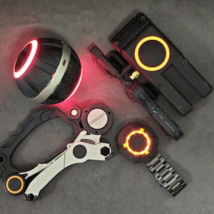 May include: A collection of futuristic electronic gadgets including a spherical device with a glowing red ring, a rectangular black module with an orange circular light, a multi-part mechanical tool with orange accents, and a circular device with an orange light.