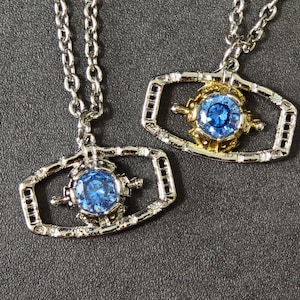 May include: Two necklaces featuring rectangular metal frames with rounded corners. Each pendant contains a central, round, faceted blue gemstone. One frame is silver-toned, while the other features a gold-toned center setting.