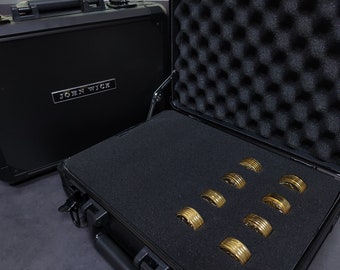 May include: A black hard-shell case with the words "JOHN WICK" on the front. The case is open and contains eight gold-colored coins inside.