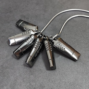 May include: A cluster of five metallic pendants, each with a cylindrical body and a rectangular top, connected to a silver chain. The pendants have a dark, gunmetal finish, and are arranged in a circular pattern against a gray background.