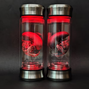 Puede incluir: Dos recipientes de vidrio con tapa y base plateadas, cada uno con una forma alienígena iluminada en rojo. Los recipientes cilíndricos están llenos de un líquido transparente. Las formas interiores son de color rojo intenso, con detalles intrincados.