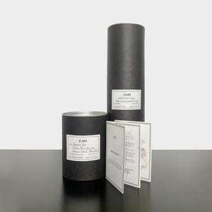 May include: Two black cylindrical boxes with white labels that read "East" and contain candles. The smaller box has a label that reads "Bergamot, Cedar, Cinnamon, Rose & Bay Leaf, Sandalwood, Patchouli, Silver Birch". The larger box has a label that reads "Hand Blended Soy & Soy Wax Organic Soy & Soy Wax Candle". There is a folded card with the text "East" and "Notes" on it.