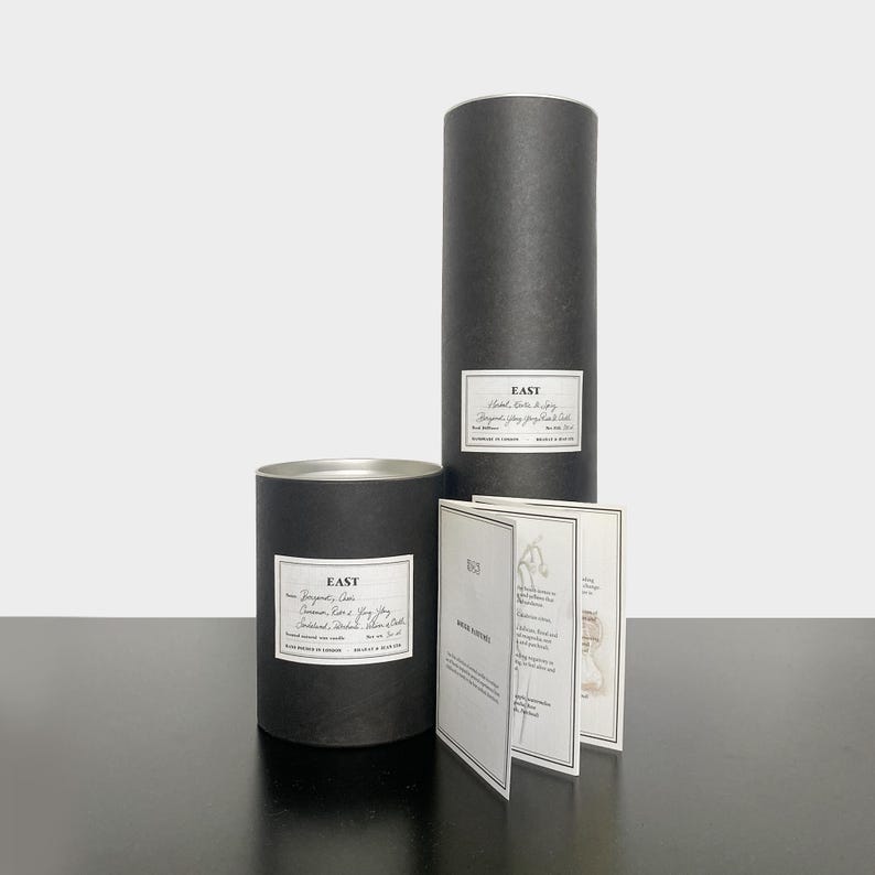 May include: Two black cylindrical boxes with white labels that read "East" and contain candles. The smaller box has a label that reads "Bergamot, Cedar, Cinnamon, Rose & Bay Leaf, Sandalwood, Patchouli, Silver Birch". The larger box has a label that reads "Hand Blended Soy & Soy Wax Organic Soy & Soy Wax Candle". There is a folded card with the text "East" and "Notes" on it.