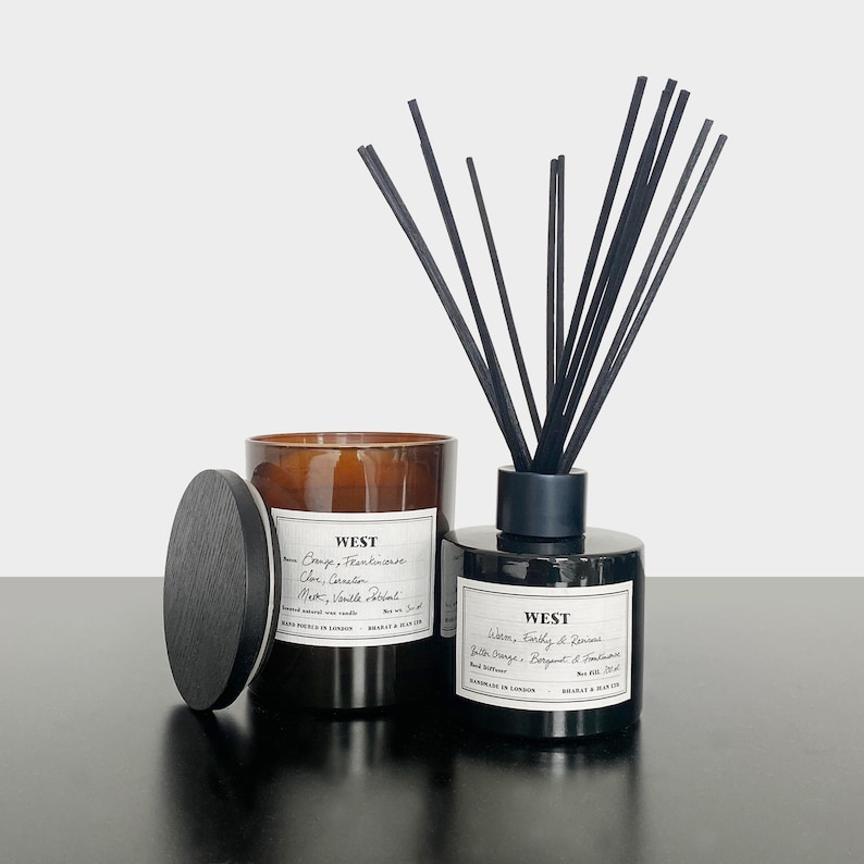 May include: Two amber glass bottles with black lids and black labels. The bottles are filled with scented oil and have black diffuser sticks. The labels read "WEST" and list the scents "Orange & Frankincense" and "Warm, Earthy & Resinous".