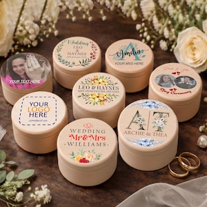 May include: A selection of personalised wooden ring boxes, each with a unique design. Some boxes display names, dates, and floral patterns, while others feature photos and the text "Your Logo Here". The boxes are circular and have a natural wood finish.