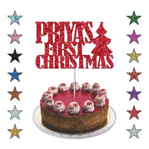 May include: A round cake with a red glaze and white frosting swirls, topped with a red glitter cake topper that reads "Priya's First Christmas" and a Christmas tree. Colourful star decorations surround the cake.