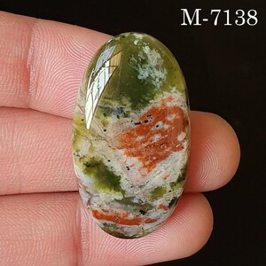May include: Oval-shaped gemstone with a mix of green, orange, and white hues. The stone has a polished surface with a speckled pattern and a reflective area. The text "M-7138" is visible in the upper right corner.