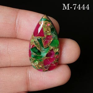 May include: A teardrop-shaped cabochon gemstone featuring a mosaic pattern of vibrant magenta and bright green fragments embedded in a metallic gold matrix. The stone is labeled with the text M-7444 in the upper right corner against a black background.