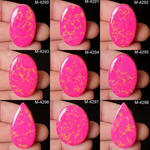 May include: Nine polished gemstones in oval and teardrop shapes, each a vivid pink with a gold matrix pattern. The stones are held in a hand, and each is labelled with a unique code. Ideal for jewellery making or collecting.