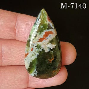 May include: A teardrop-shaped gemstone with a polished surface. The stone features a mix of green, orange, and white patterns. The text "M-7140" is visible in the upper right corner. The gemstone is held by a person.