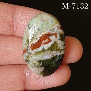 May include: An oval-shaped gemstone with a mix of colors, including green, red, and white. The stone has a polished surface and a natural, organic appearance. The text "M-7132" is visible in the upper right corner.