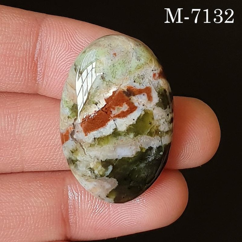 May include: An oval-shaped gemstone with a mix of colors, including green, red, and white. The stone has a polished surface and a natural, organic appearance. The text "M-7132" is visible in the upper right corner.