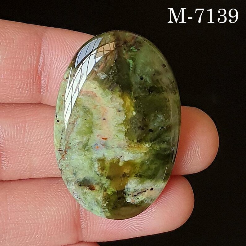 May include: An oval-shaped gemstone with a polished surface. The stone features a blend of green, brown, and yellow hues with dark speckles. The text "M-7139" is visible in the upper right corner. The gemstone is held between fingers.