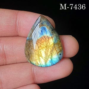 Pode incluir: Uma pedra preciosa de labradorite em forma de gota com uma superfície facetada com reflexos iridescentes azuis e dourados, exibindo um padrão interno rachado distinto e o texto M-7436 impresso no canto superior direito da imagem.