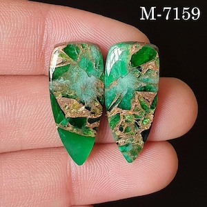Pode incluir: Duas pedras preciosas em forma de gota, com um tom verde vibrante, entremeadas por veios dourados e castanhos. As pedras estão sobre um fundo escuro. O texto "M-7159" é visível no canto superior direito.