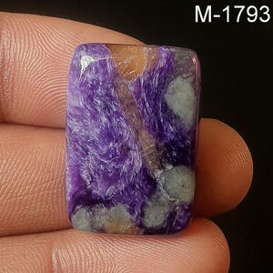 Puede incluir: Una piedra preciosa cabujón de color púrpura, gris y naranja con un patrón de remolino fibroso. La piedra mide aproximadamente 5 cm de largo y 4 cm de ancho.