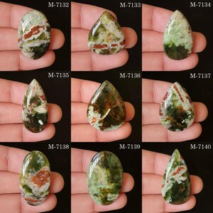 May include: Nine polished gemstones in various shapes, held in a hand. The stones display a mix of green, brown, and white colors. Each stone has a unique pattern and is labeled with a number from M-7132 to M-7140.