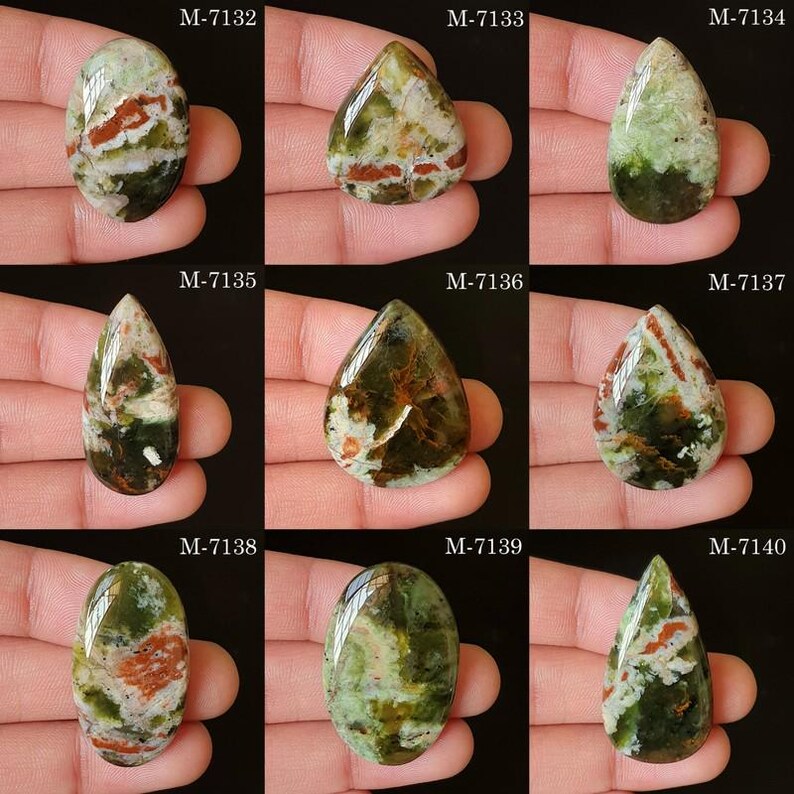 May include: Nine polished gemstones in various shapes, held in a hand. The stones display a mix of green, brown, and white colors. Each stone has a unique pattern and is labeled with a number from M-7132 to M-7140.