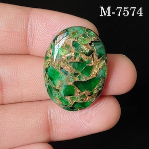 Puede incluir: Una piedra preciosa cabujón de forma ovalada con un patrón de mosaico verde vibrante y vetas de color cobre metálico en toda la superficie. La piedra está marcada con el texto M-7574 en la esquina superior derecha sobre un fondo oscuro.