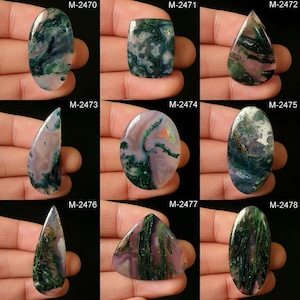 Puede incluir: Nueve cabujones de ágata verde y rosa diferentes, cada uno con un patrón y una forma únicos. Los cabujones se muestran en una mano, con el código del producto etiquetado encima de cada piedra.