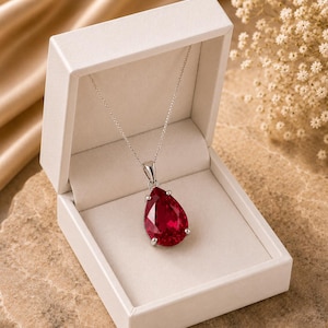 Può includere: Una collana in argento con un grande ciondolo a forma di pera in pietra preziosa rosso intenso, fissato da una montatura a quattro griffe. La collana è esposta all'interno di un portagioie rettangolare aperto color crema con finitura opaca.