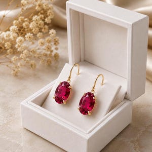 May include: A pair of oval-shaped, deep pink gemstone drop earrings set in gold metal. The earrings feature a faceted cut and are displayed inside an open, square-shaped white jewelry box with a hinged lid and a soft, padded interior lining.
