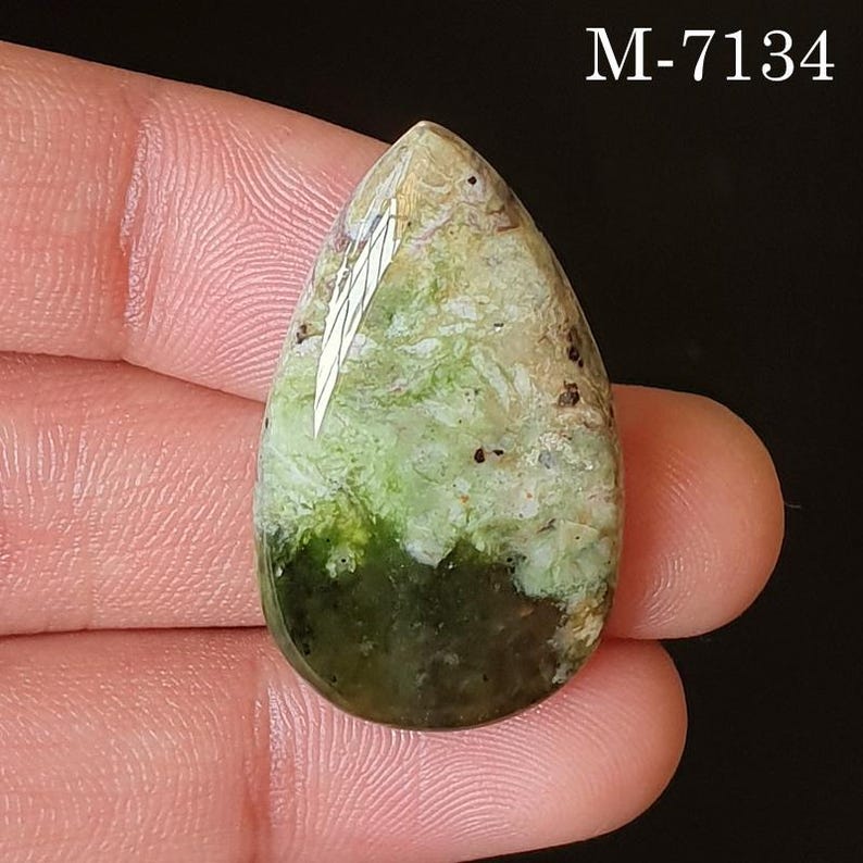 May include: A teardrop-shaped gemstone with a mix of green, beige, and brown hues. The stone has a polished surface with a gradient of colors, from dark green at the base to lighter shades at the top. The text "M-7134" is visible.