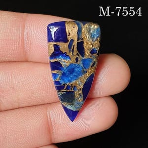 Puede incluir: Un cabujón de piedra preciosa pulida de forma triangular que presenta una base azul real profundo con intrincadas vetas de color oro metálico en toda la superficie. El texto M-7554 está impreso en blanco en la esquina superior derecha.
