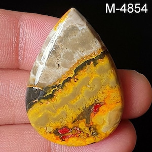May include: A teardrop-shaped gemstone with a unique pattern of yellow, orange, black, and tan colors. The stone has a polished surface and a natural, organic appearance. The text "M-4854" is visible in the upper right corner. This gemstone is ideal for jewelry making.