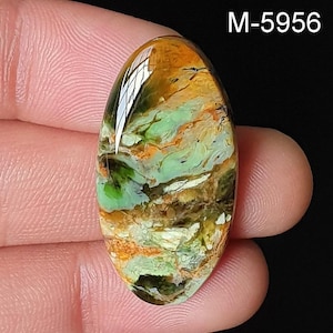 May include: An oval gemstone with a polished surface. The stone displays a mix of green, orange, brown, and white hues, creating a marbled effect. The text "M-5956" is visible. The gemstone is held between fingers. The stone is likely for jewellery.