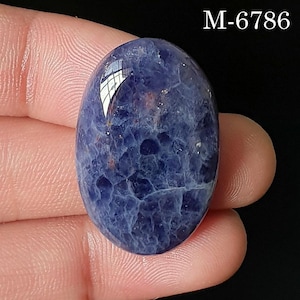 Peut inclure: Une pierre précieuse bleue polie de forme ovale avec une surface texturée et marbrée présentant des veines bleu indigo profond et bleu plus clair. La pierre a une finition brillante et porte le texte M-6786 dans le coin supérieur droit.