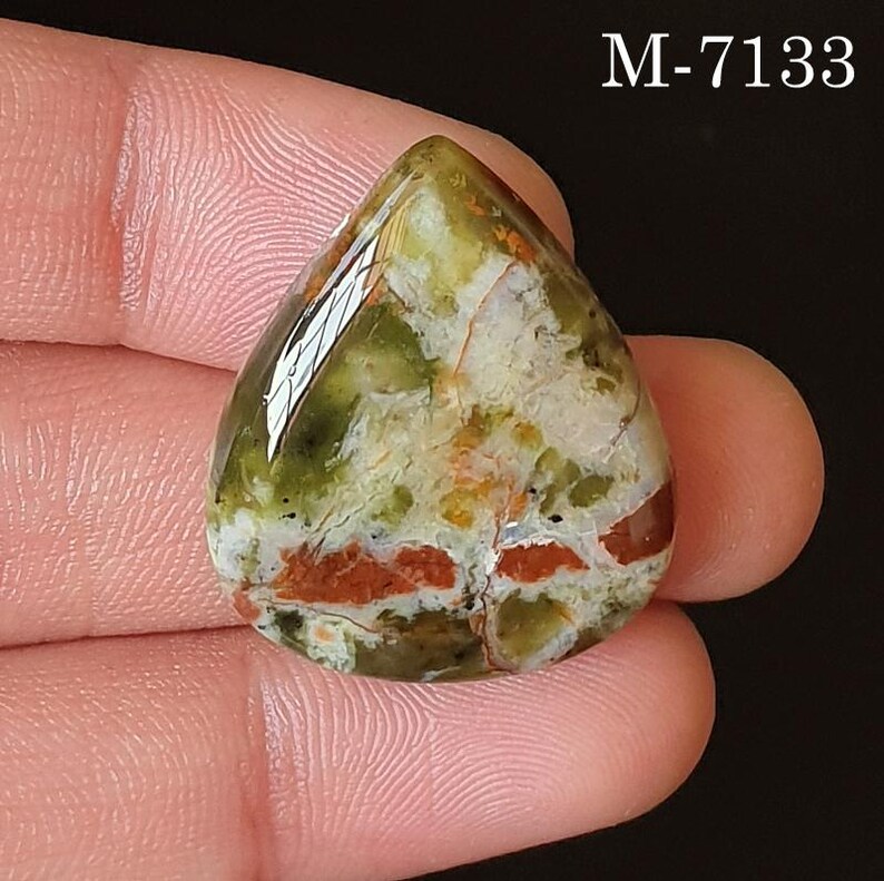 May include: A teardrop-shaped gemstone with a polished surface. The stone features a mix of colors, including green, white, and red-brown, with a glossy finish. The text "M-7133" is visible in the upper right corner. The gemstone is held in a hand.