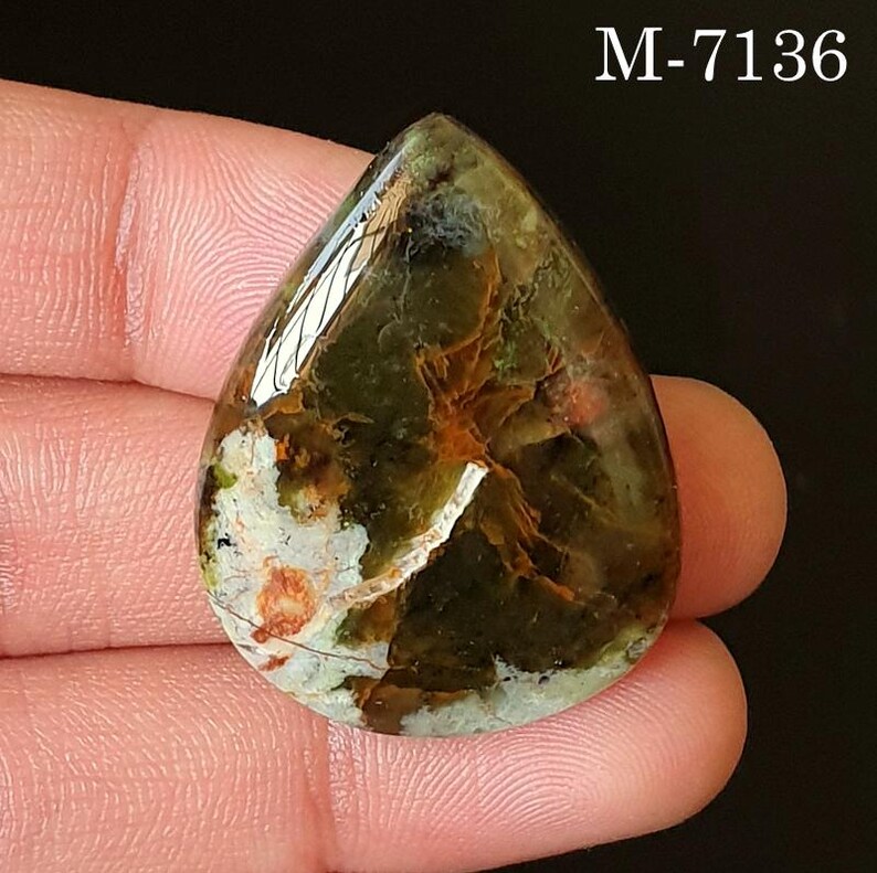 May include: A teardrop-shaped gemstone with a polished surface. The stone features a mix of green, brown, white, and orange hues. The text "M-7136" is visible in the upper right corner. The gemstone is held between fingers.