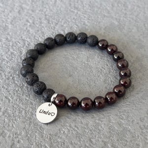 Peut inclure: Un bracelet de perles composé de perles de roche volcanique gris foncé et de perles de grenat rouge foncé. Un petit charme rond en argent avec le nom "Linda" et un cœur est attaché.