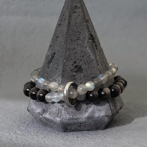 Könnte beinhalten: Zwei Armbänder mit Perlen liegen auf einem dunkelgrauen, geometrischen Präsentationsständer. Ein Armband hat schwarze Perlen, das andere graue und irisierende Perlen. Zwischen den Armbändern befindet sich ein silberner Ring.