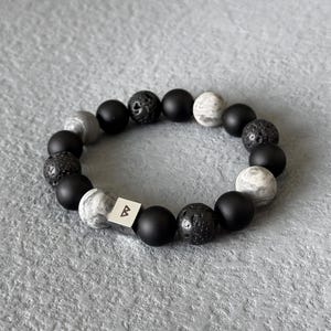 Op de afbeelding: Een kralenarmband met zwarte, grijze en witte stenen kralen. De armband heeft matte zwarte kralen, lavasteenkralen en grijze en witte kralen met patronen. Een zilveren kubuskraal met de letter "B" is ook inbegrepen.