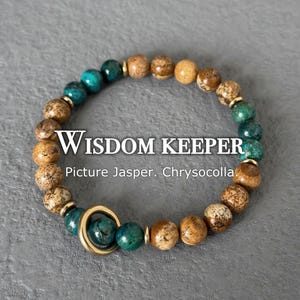 May include: A beaded bracelet featuring Picture Jasper and Chrysocolla stones. The bracelet has a mix of brown, tan, and teal beads, with gold-colored accents. The text "Wisdom Keeper" is displayed on the bracelet.