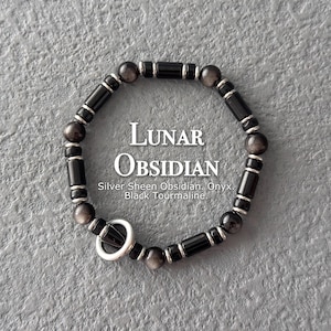 May include: A black bracelet with silver detailing, composed of beads and cylindrical onyx elements. The bracelet has a silver ring clasp. The image text reads "Lunar Obsidian" and lists materials: Silver Sheen Obsidian, Onyx, Black Tourmaline.