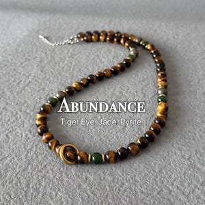 May include: A beaded necklace featuring tiger eye, jade, and pyrite stones. The necklace has a gold-colored clasp and a silver chain. The word "ABUNDANCE" is displayed in white text, along with the stone names.