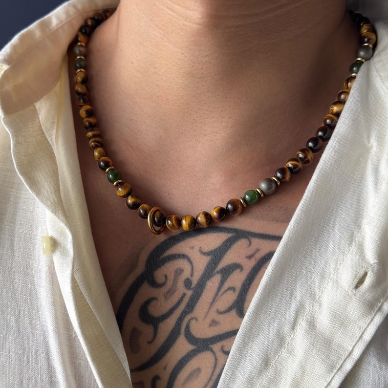 May include: A beaded necklace featuring tiger eye stones, interspersed with green and gray beads. The necklace has a gold-colored clasp and is worn around the neck. The necklace is a piece of jewelry.