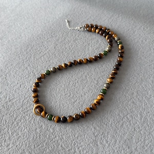 May include: A beaded necklace featuring tiger's eye, green, and metallic beads. The necklace has a gold-colored ring clasp and a silver chain. The beads are strung together to create a stylish accessory.