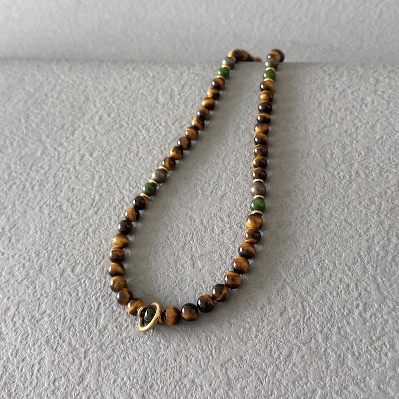 May include: A beaded necklace featuring tiger's eye beads, interspersed with green and gray beads, and gold-colored accents. The necklace has a gold-colored clasp. The beads are approximately 0.3 inches in diameter.