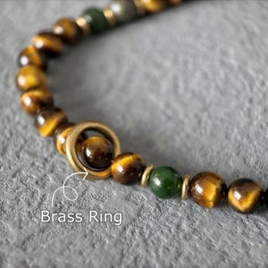 May include: Close-up of a beaded bracelet featuring tiger eye and green beads, with a brass ring accent. The beads are a mix of brown and gold, with a polished finish. The text "Brass Ring" is visible.