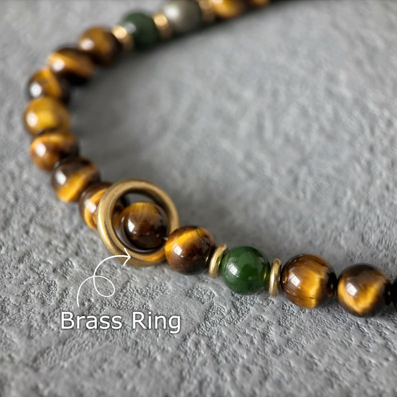 May include: Close-up of a beaded bracelet featuring tiger eye and green beads, with a brass ring accent. The beads are a mix of brown and gold, with a polished finish. The text "Brass Ring" is visible.
