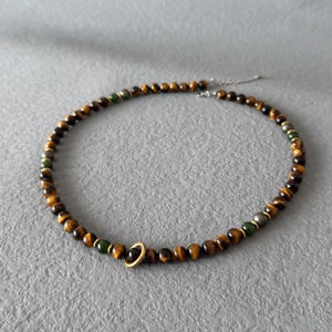May include: A beaded necklace featuring tiger eye and green beads, with a gold-colored ring pendant. The necklace is strung on a silver chain. The beads are approximately 0.25 inches in diameter.