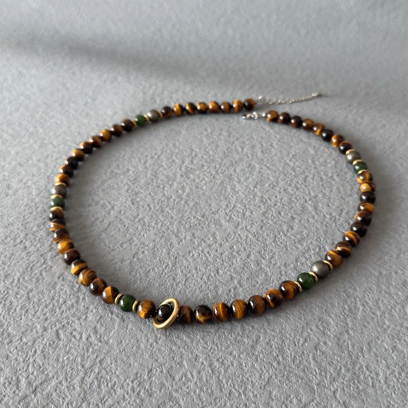 May include: A beaded necklace featuring tiger eye and green beads, with a gold-colored ring pendant. The necklace is strung on a silver chain. The beads are approximately 0.25 inches in diameter.