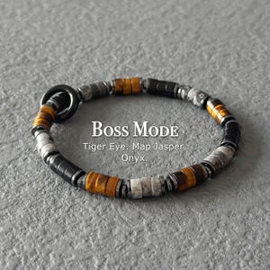 Könnte beinhalten: Ein Perlenarmband mit zylindrischen Tigerauge-, Landschaftsjaspis- und Onyxperlen in einem sich wiederholenden Muster. Das Armband hat einen schwarzen runden Verschluss und zeigt in der Mitte den Text BOSS MODE Tiger Eye. Map Jasper. Onyx.
